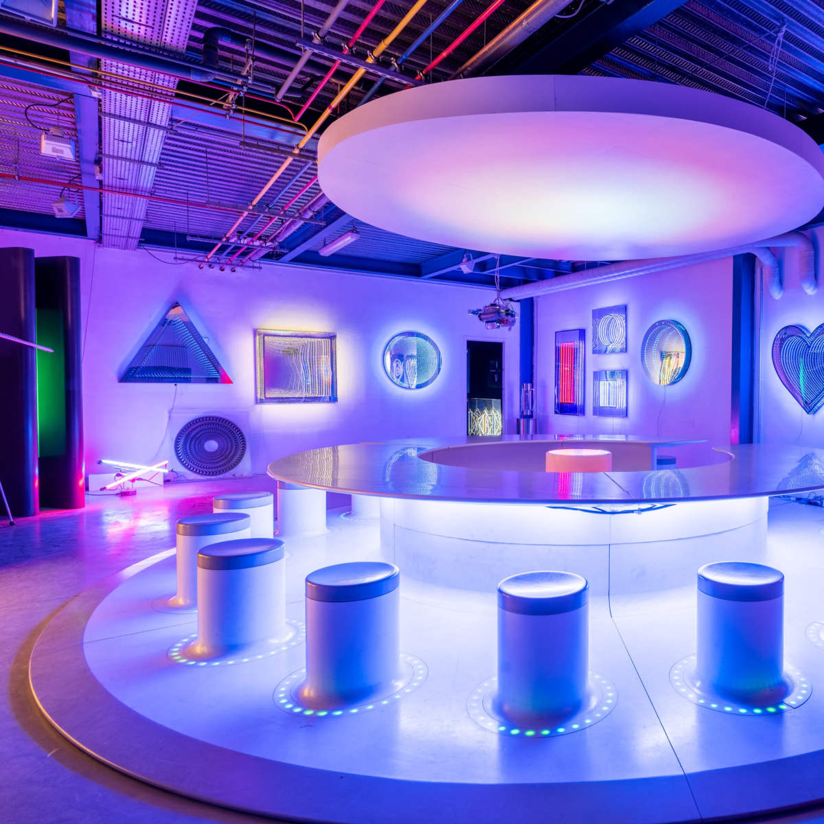 A modern, vibrant lounge space featuring a circular table surrounded by high stools, illuminated by colorful neon lights and art installations on the walls.