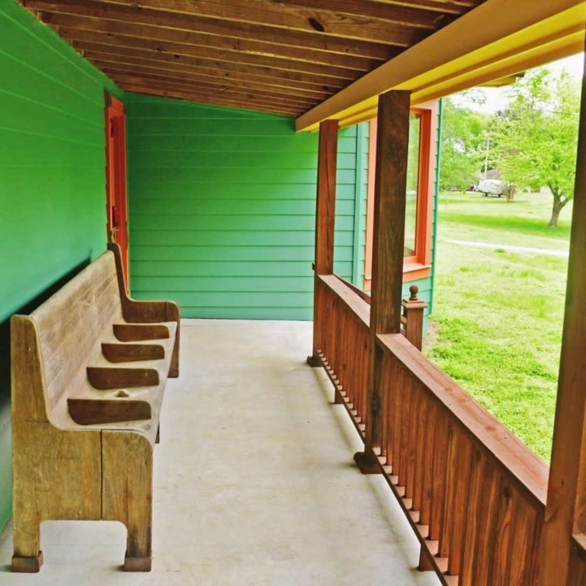 A green-painted porch with a wooden bench and a view of a grassy area outside.