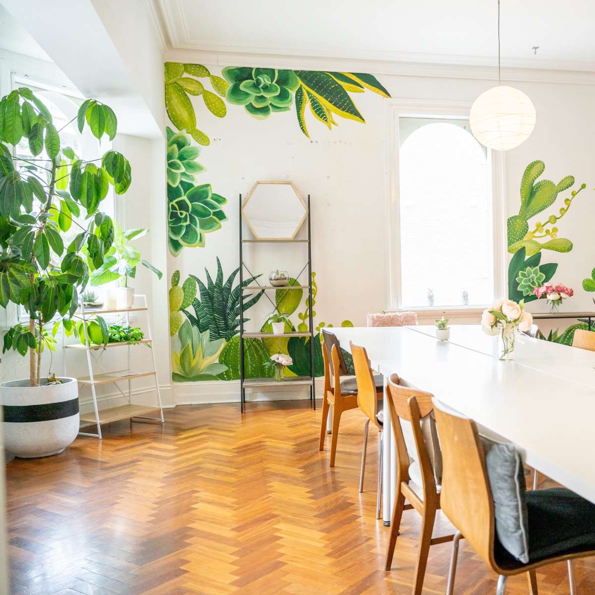 A bright dining area featuring a large white table surrounded by wooden chairs, with green plants and a botanical mural on the wall.