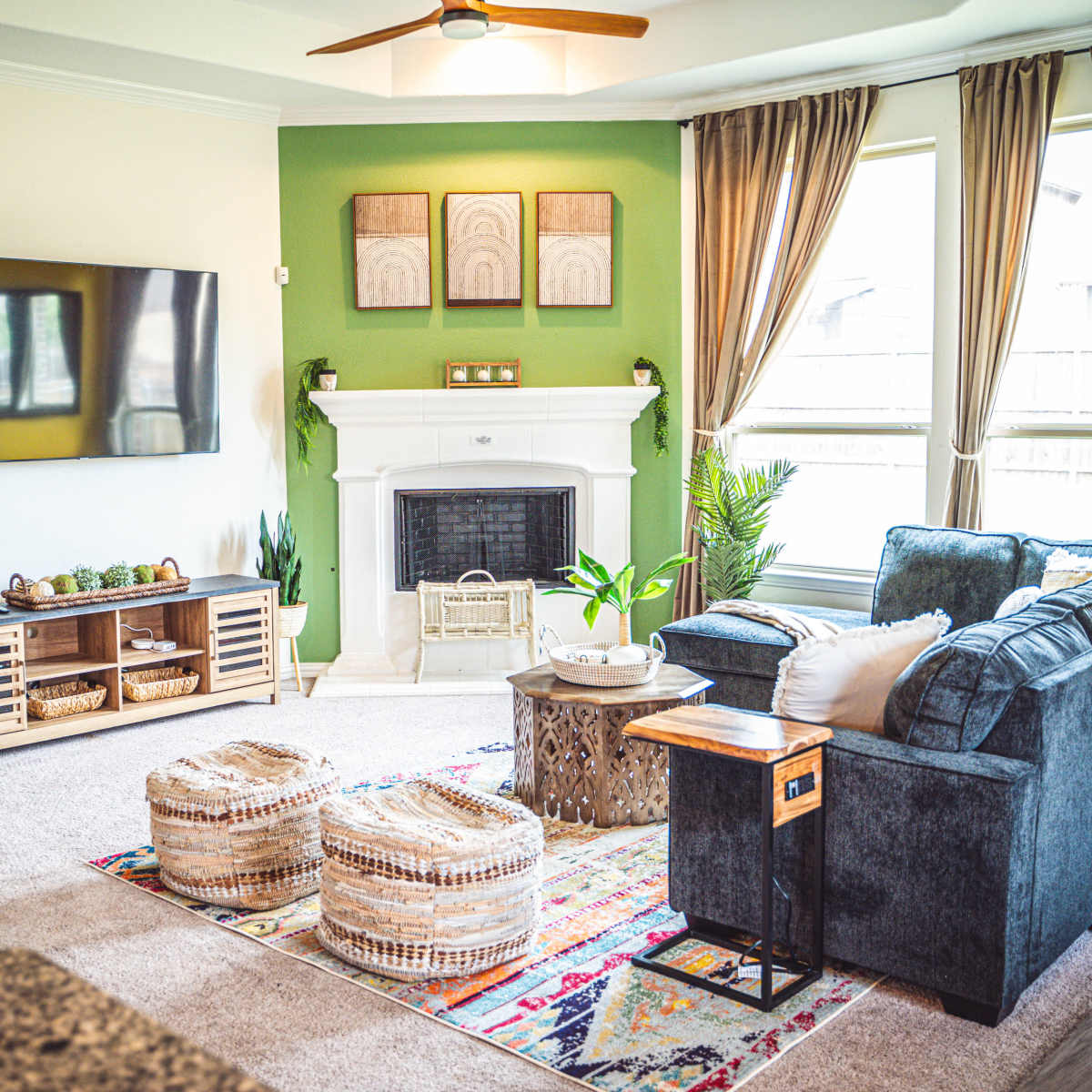 The living room features a television mounted on the wall, a fireplace with decorative items, and a seating area with a sofa and two woven stools.