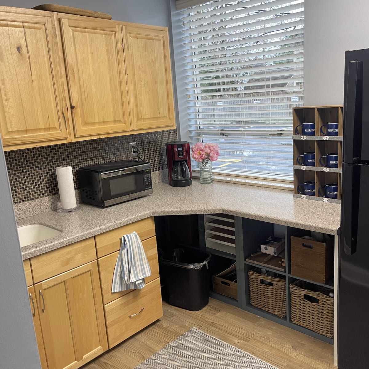 A compact kitchen features wooden cabinets, a countertop with a sink, a microwave, a coffee maker, and a black refrigerator, alongside a window with blinds and decorative shelving.