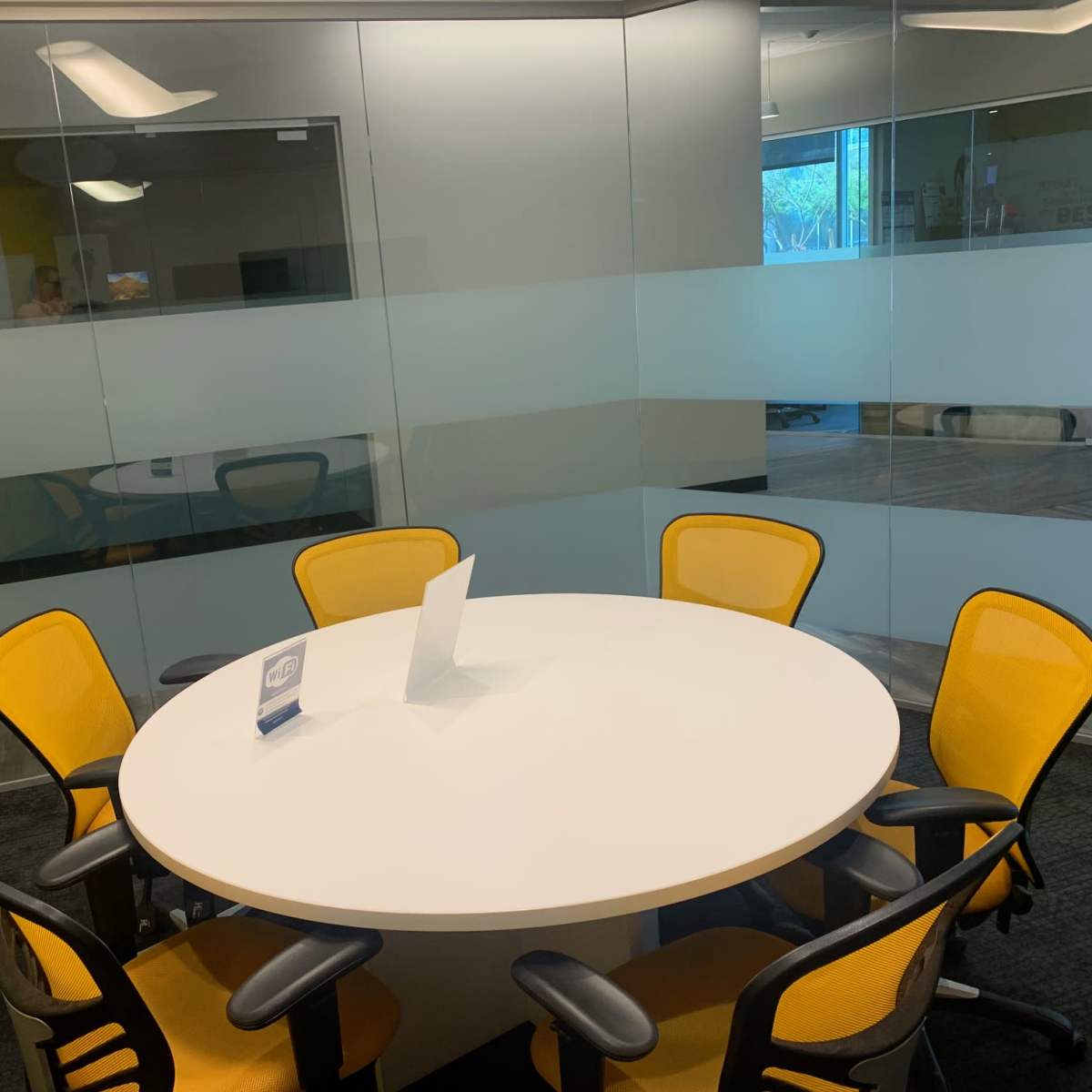 A round table with a white surface is surrounded by six yellow chairs in a glass-walled meeting room.
