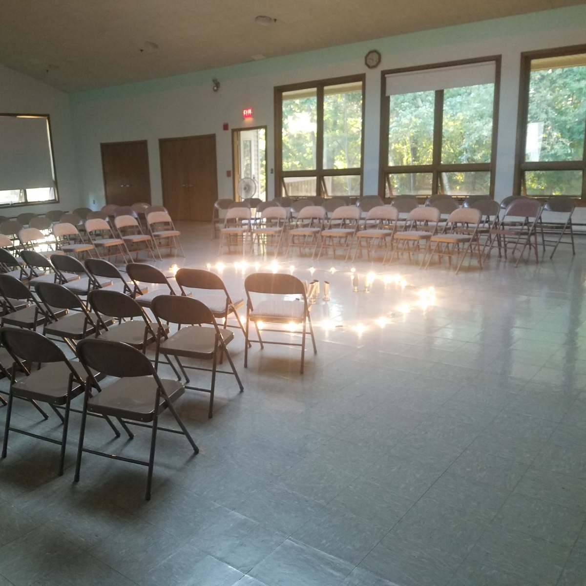 A room is arranged with rows of folding chairs facing a circular formation in the center, illuminated by natural light from large windows.