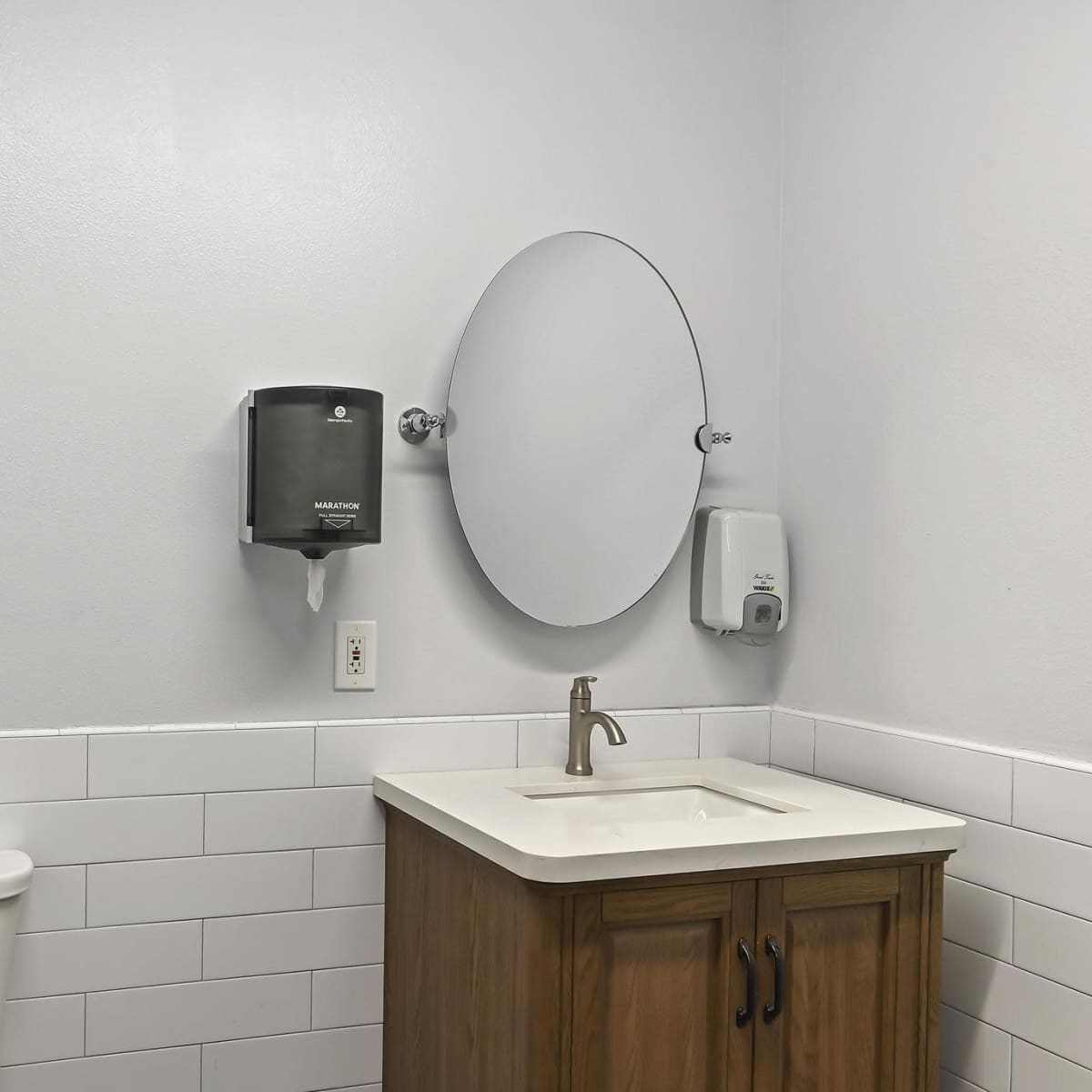 A clean bathroom featuring a toilet, a sink with a wooden cabinet, a mirror, and a soap dispenser on the wall.