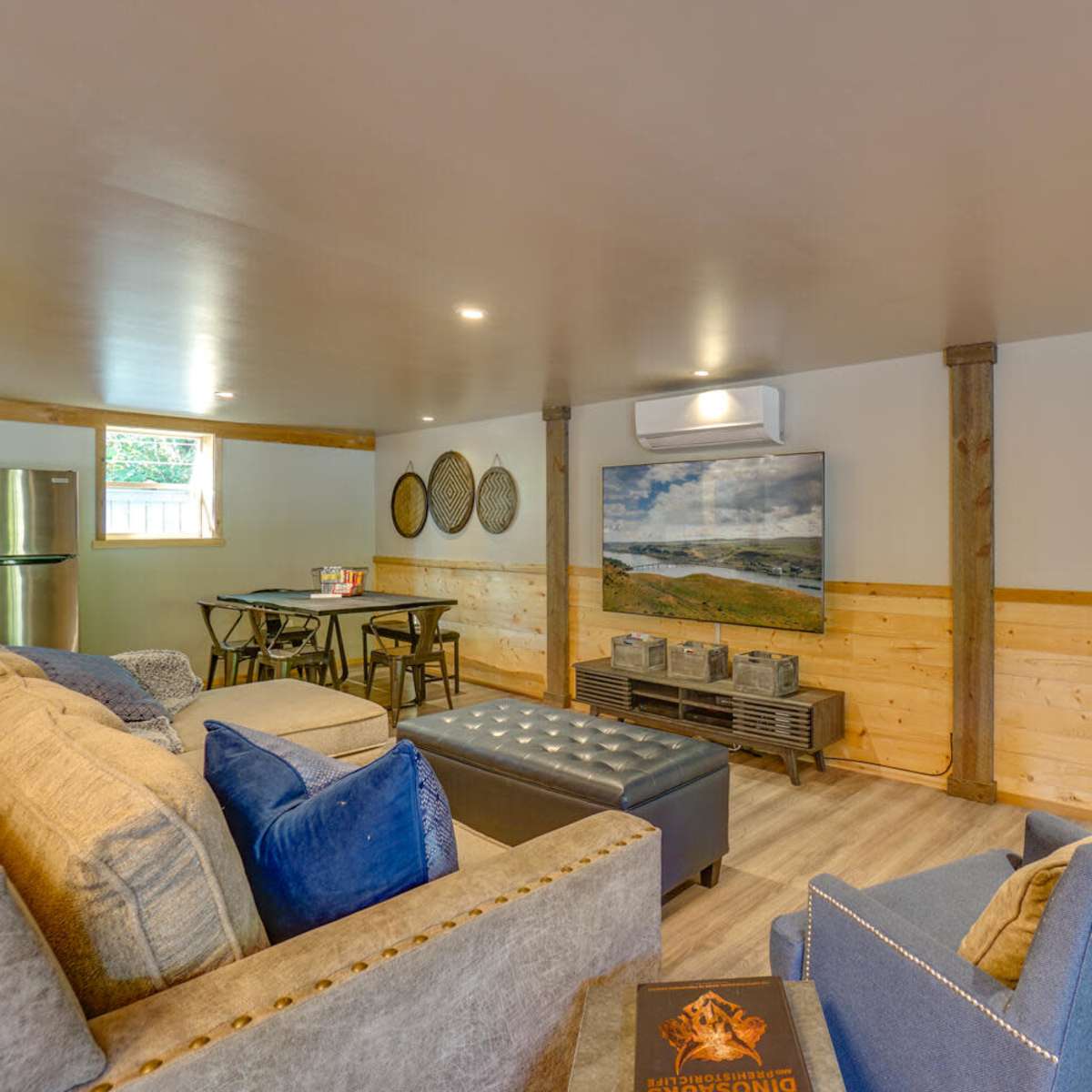 A cozy basement living area with a sofa, a dining table, and a wall-mounted TV, featuring wood paneling and modern decor.