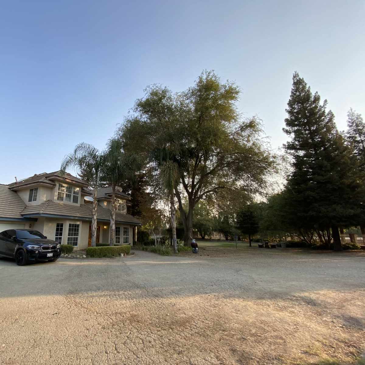 A two-story house with a tiled roof and a black car parked in the driveway is surrounded by trees and a gravel pathway.