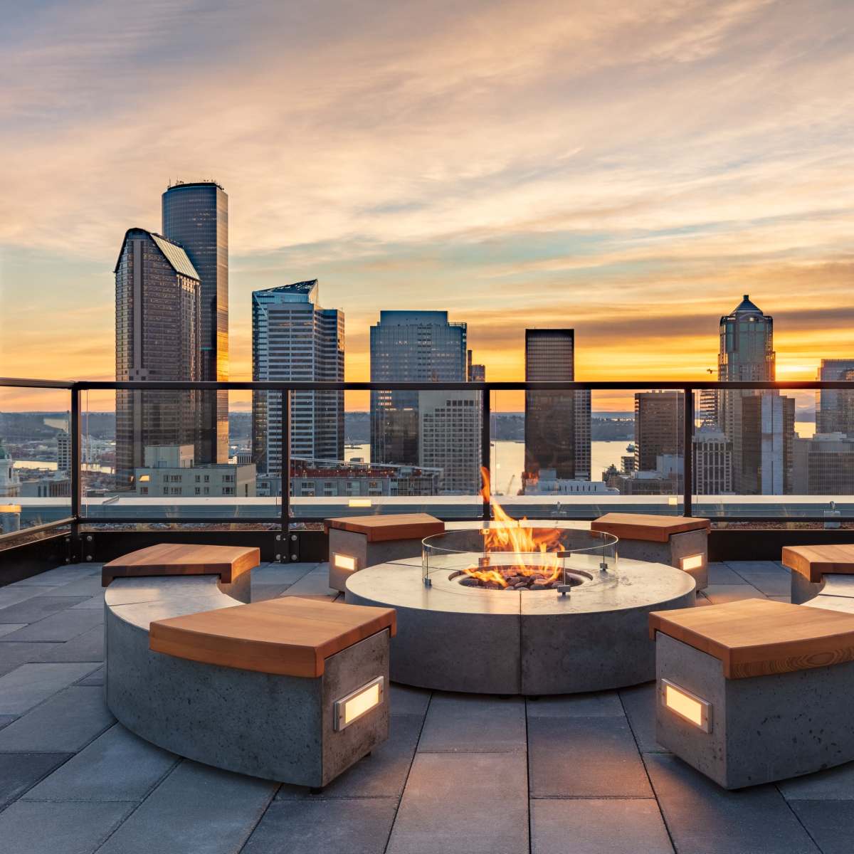 A rooftop lounge features a circular seating arrangement around a central fire pit, with city skyscrapers and a sunset in the background.