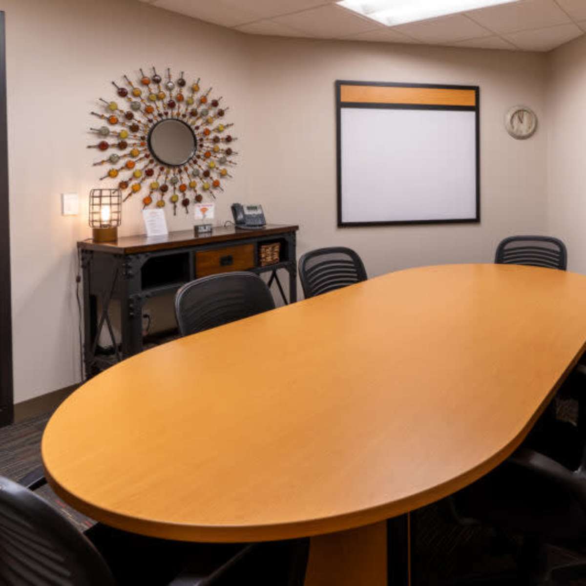 A conference room features an oval wooden table surrounded by black chairs, with a circular decorative mirror on one wall and a blank whiteboard on another.