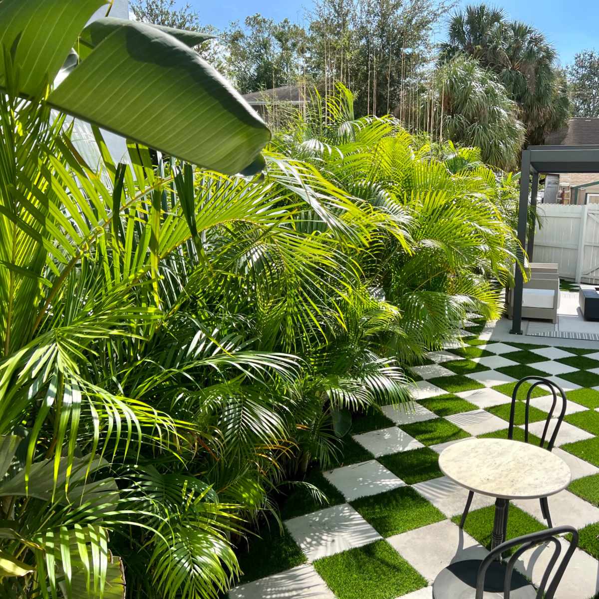 A small patio features a round marble table and black chairs, surrounded by lush greenery and a checkerboard pattern of artificial grass and stone tiles.