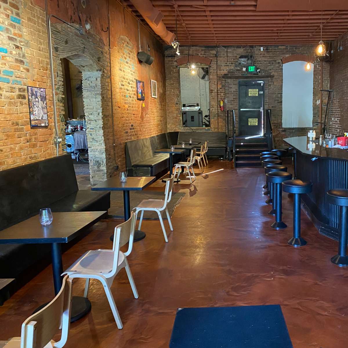 A dimly lit restaurant interior with brick walls, a bar on the right, and several tables and chairs lining the left side.