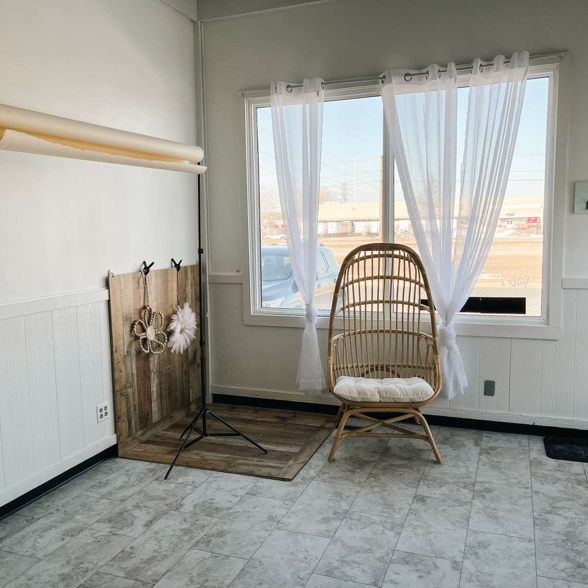 A brightly lit room features a rattan chair next to a window, with sheer curtains and a wooden backdrop.
