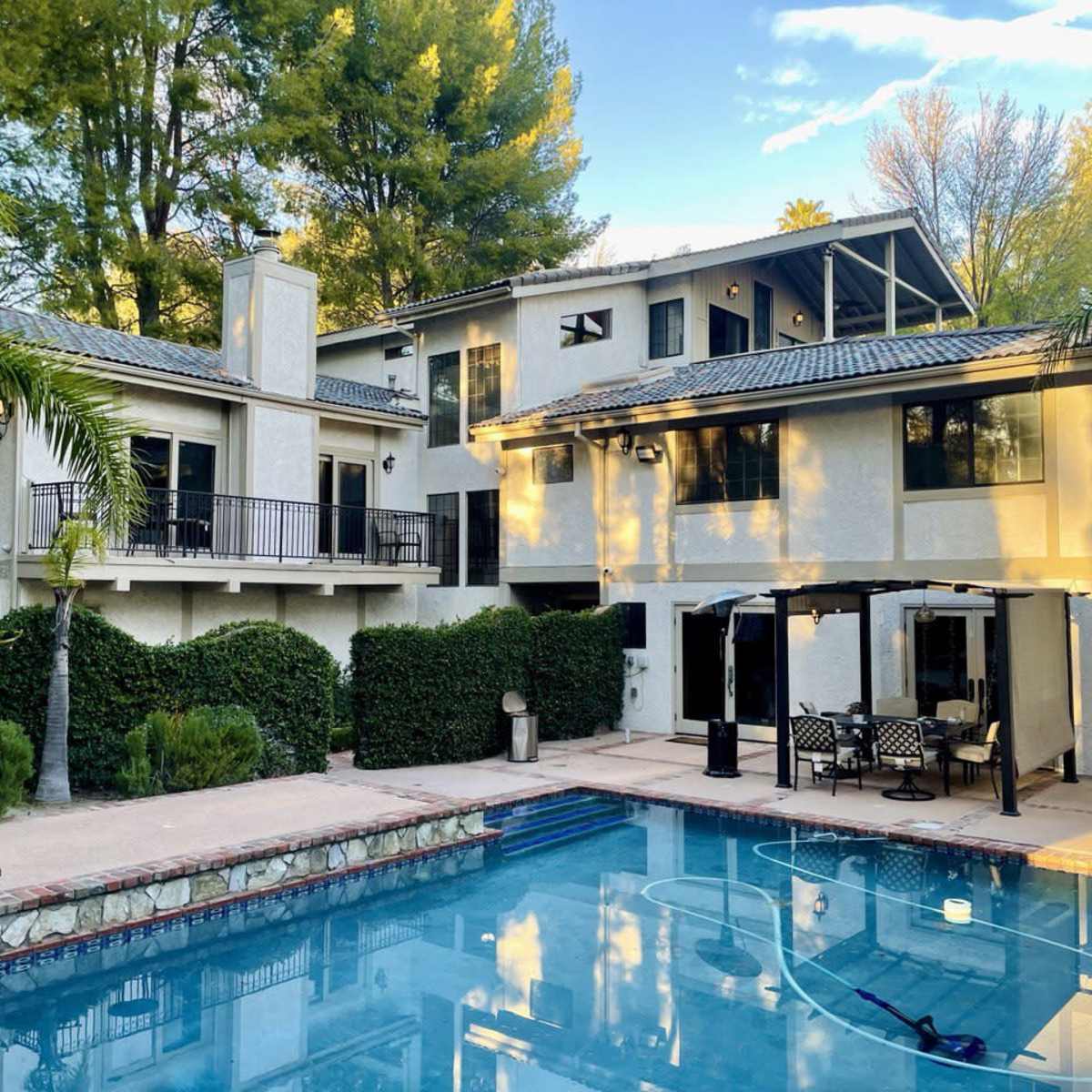 A two-story house adjacent to a swimming pool surrounded by trees and a patio area with outdoor furniture.