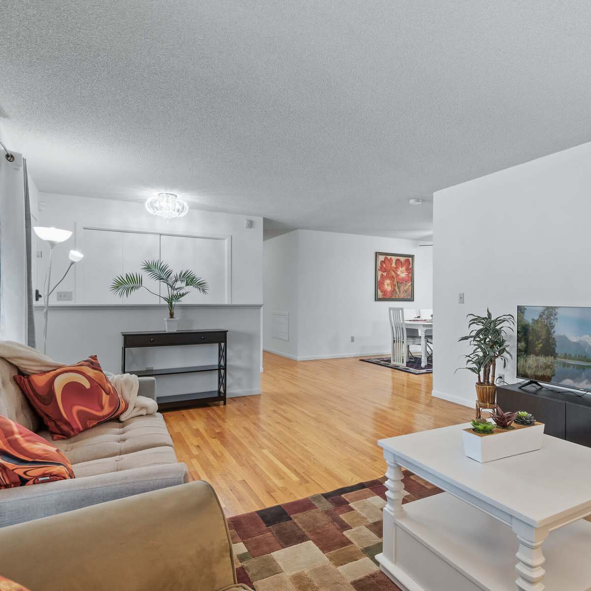 A modern living room features a beige sofa with colorful cushions, a white coffee table, a television on a stand, and wooden flooring.