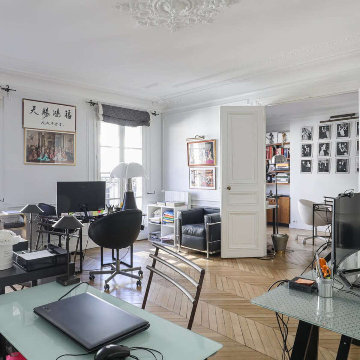 A modern office space features multiple workstations, a variety of chairs, and a separation between two rooms, with art displayed on the walls.