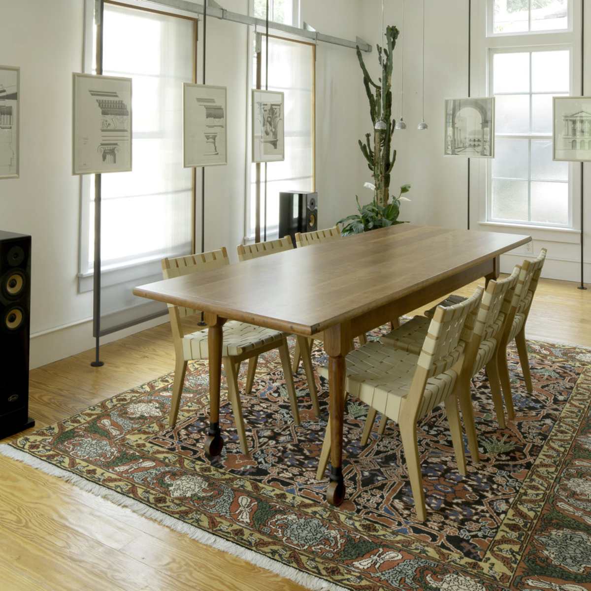A wooden dining table surrounded by six chairs sits on a patterned rug in a well-lit room with large windows and hanging artwork.