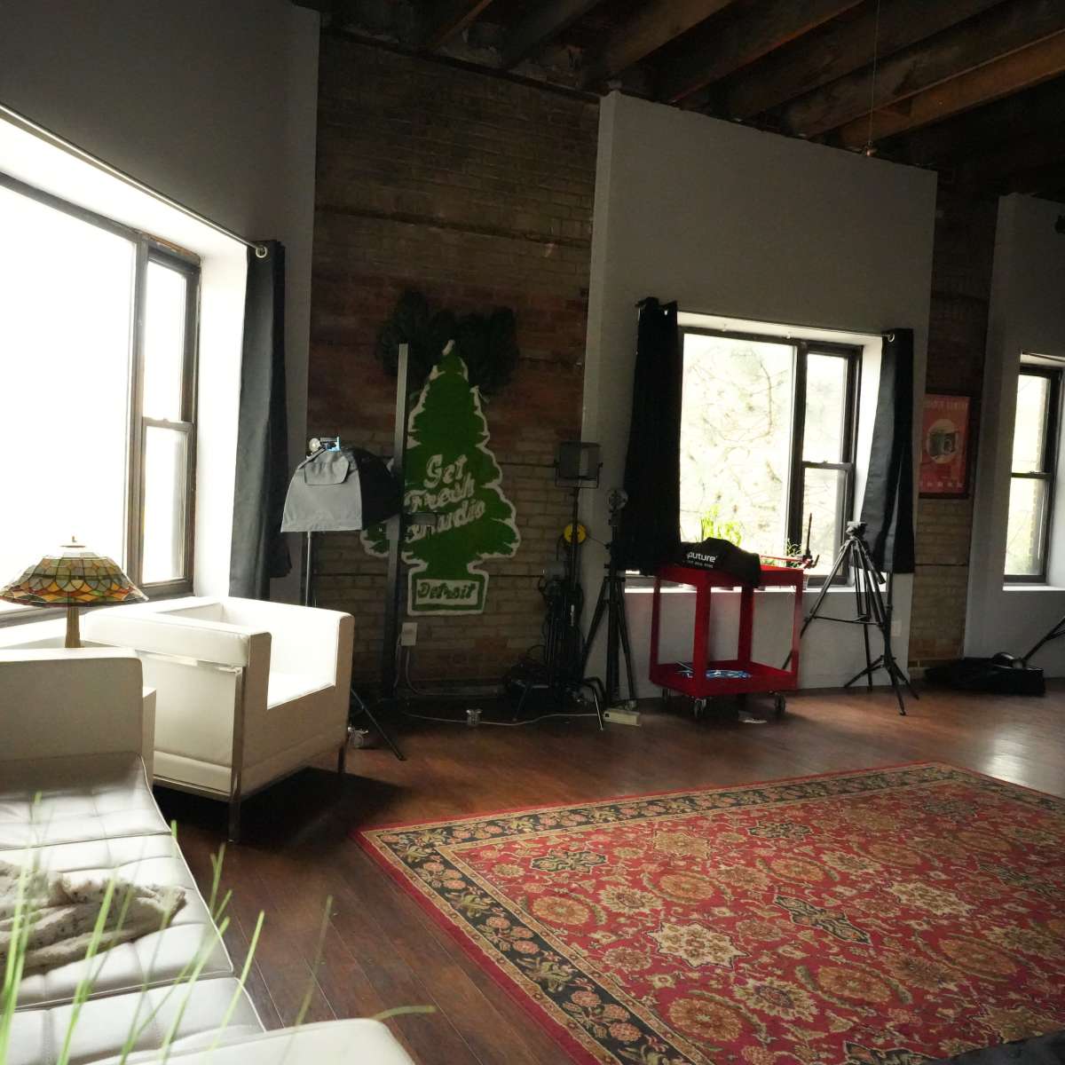 A spacious room with large windows, a white leather sofa, a patterned rug, and various lighting equipment in the background.