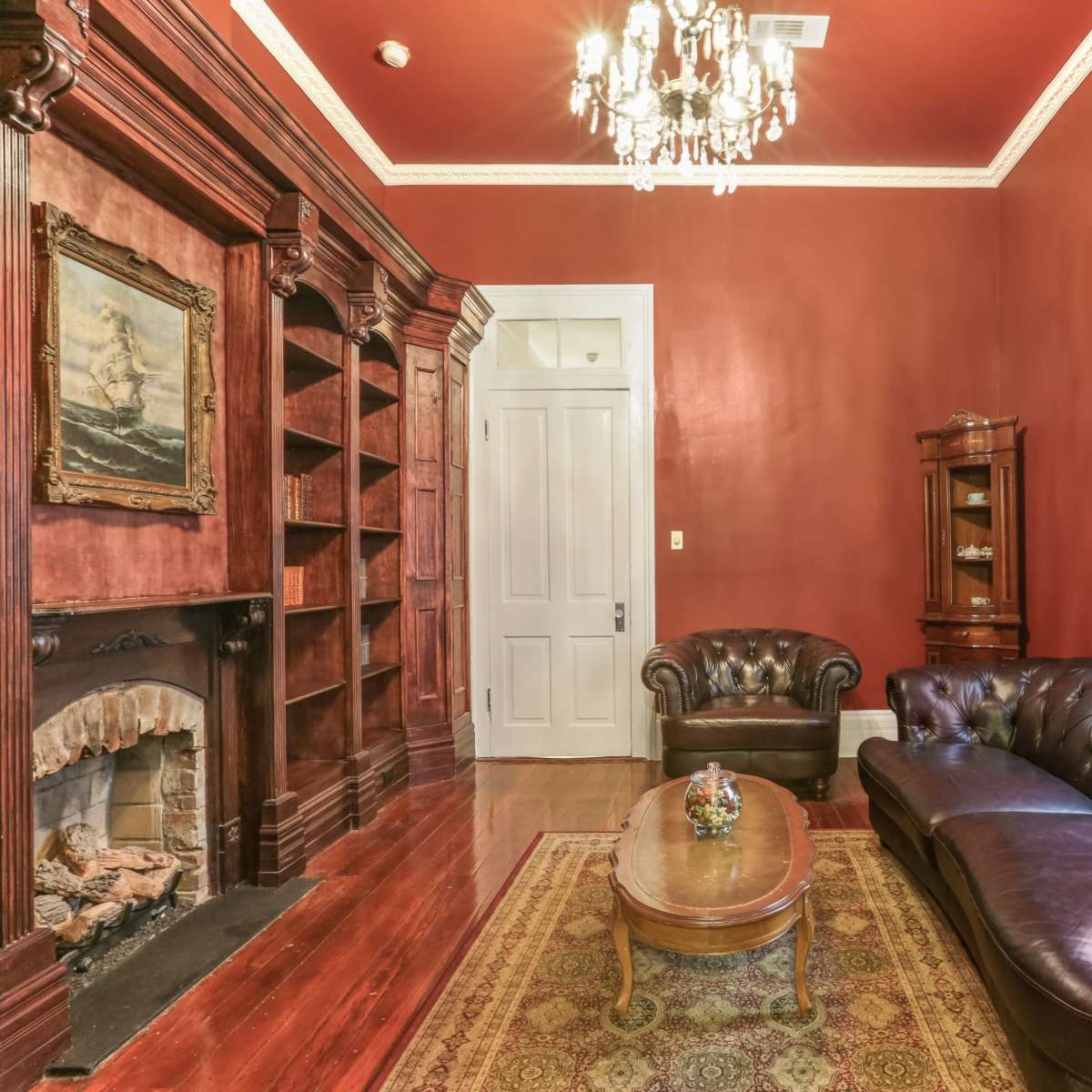 A warmly decorated room with a dark red wall, a leather sofa, a coffee table, and bookshelves flanking a fireplace.