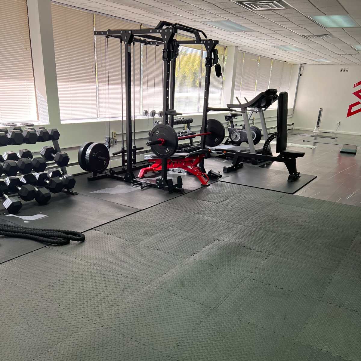 A gym interior with weightlifting equipment, including a squat rack and dumbbells, arranged on a rubber mat flooring.