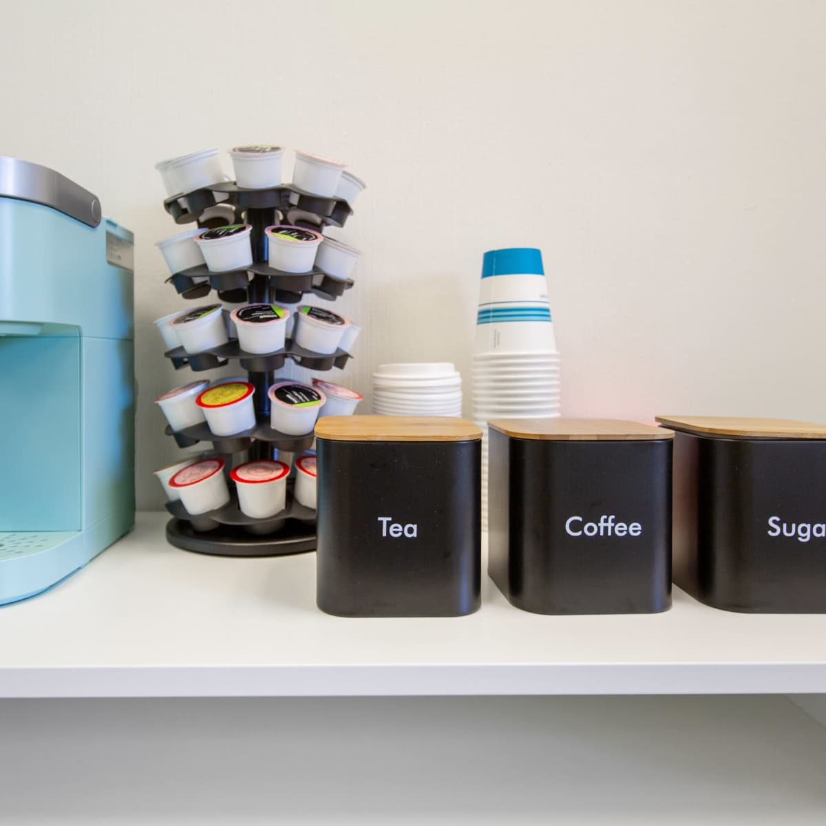 A light blue Keurig coffee maker, a vertical carousel of coffee pods, and three black containers labeled "Tea," "Coffee," and "Sugar" on a white shelf.