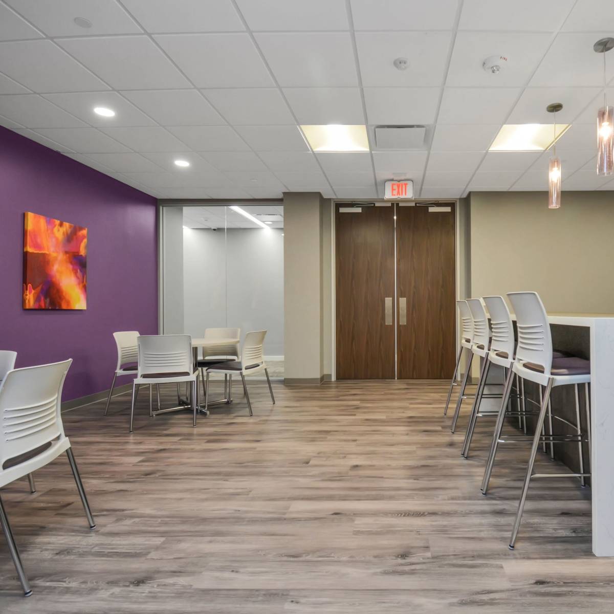 A modern break room with a purple accent wall, a kitchenette area, and several white tables and chairs.