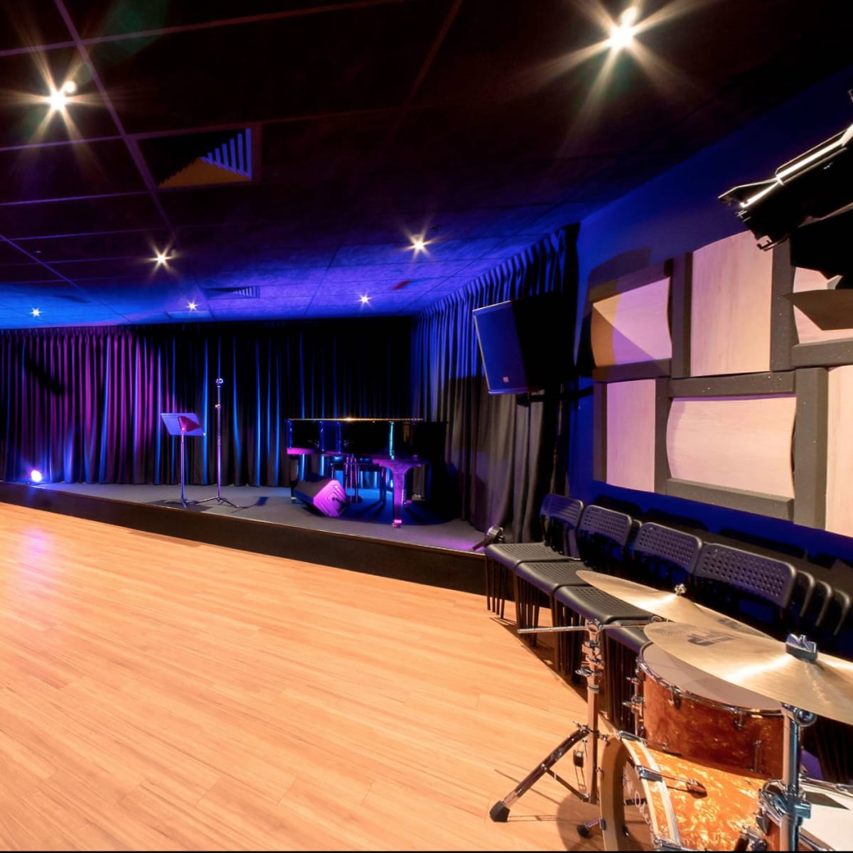 A music studio featuring a stage area with a microphone, a piano, a drum set, and seating along the sidewalls, all under soft stage lighting.