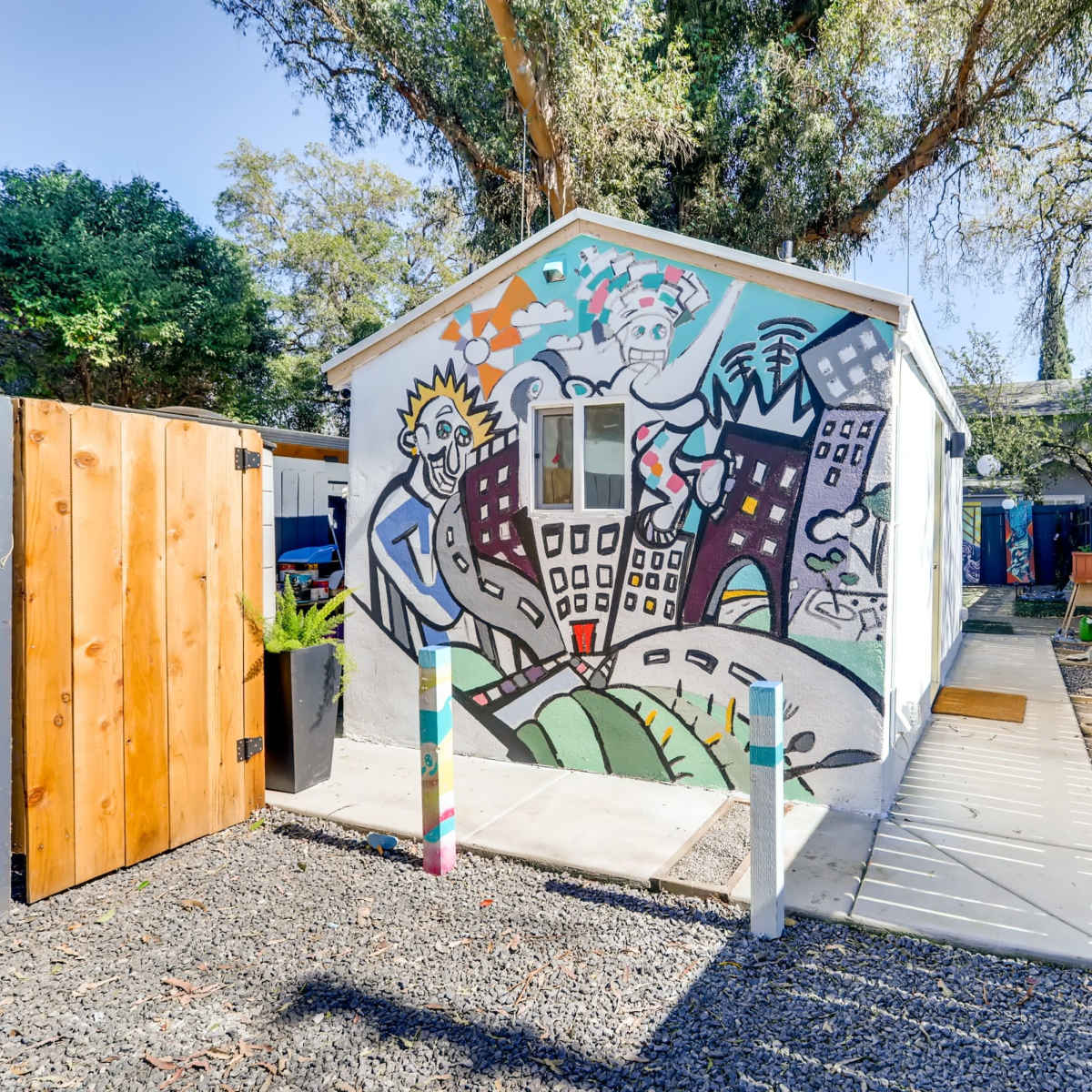 The scene depicts a colorful mural on a small building surrounded by a gravel pathway and landscaping elements in a garden setting.