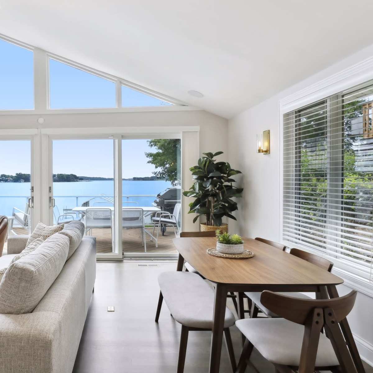 A bright, modern living area features a large window overlooking a lake, with comfortable seating and a dining table nearby.