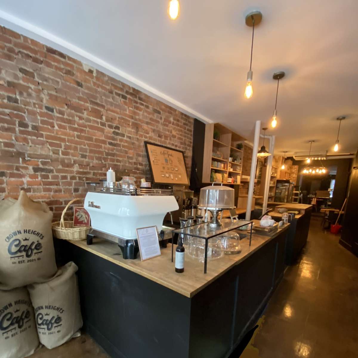 A cozy coffee shop interior featuring a brick wall, a wooden counter with coffee equipment, and bags of coffee beans in the foreground.