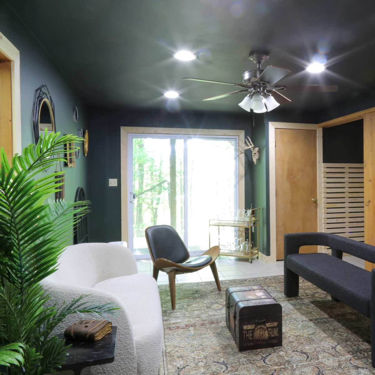 The room features a modern seating arrangement with a light-colored sofa, a black bench, and a chair, illuminated by ceiling fans and natural light from sliding doors that lead to an outdoor area.