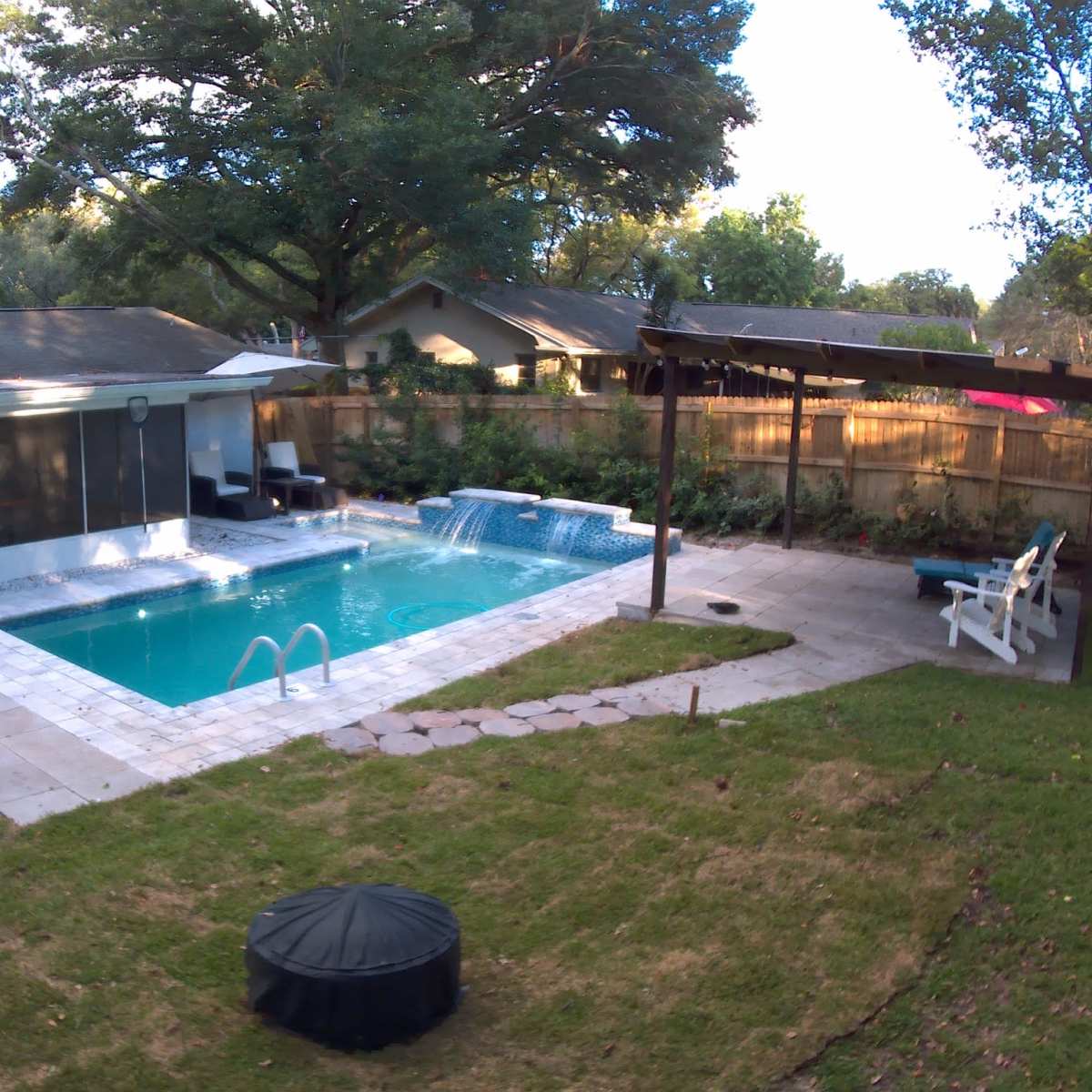 A backyard with a swimming pool, a covered patio area, and a lawn surrounded by trees and decorative landscaping.