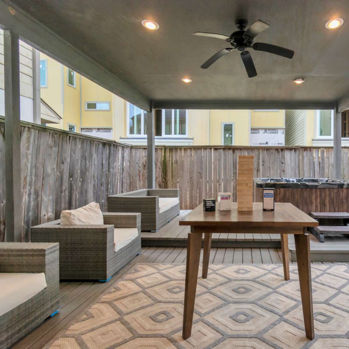 A covered patio features a wooden table, two seating areas, and a hot tub surrounded by wooden fencing.