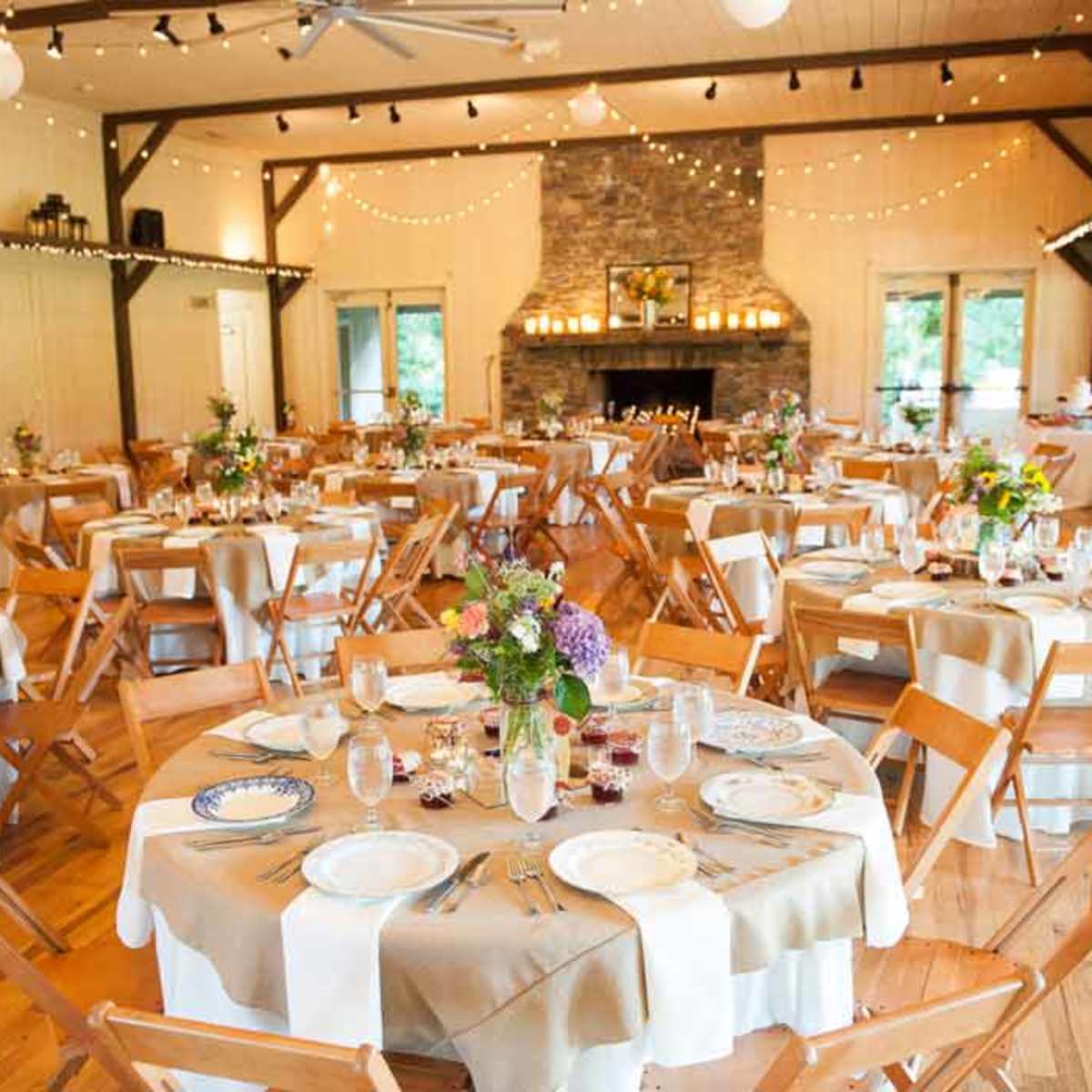 A bright banquet hall set for a formal event, featuring round tables adorned with floral centerpieces and accompanied by wooden chairs.