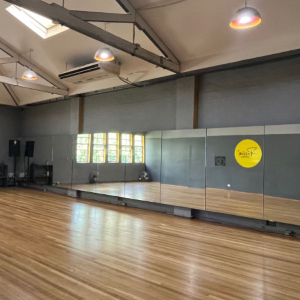 A spacious dance studio features a wooden floor, large mirrors along one wall, and several windows allowing natural light.