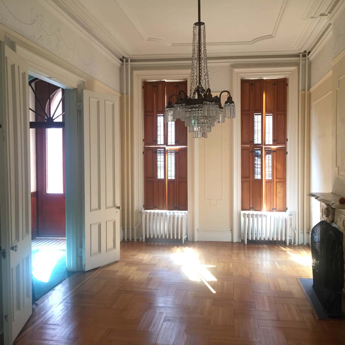 A spacious room with wooden floors, tall windows, and a chandelier, featuring doorways leading to another area.