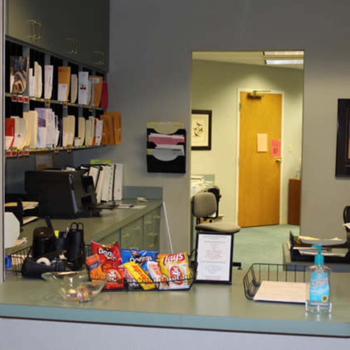 An office reception area features a counter with snacks and a hand sanitizer, while a hallway leads to an office space in the background.