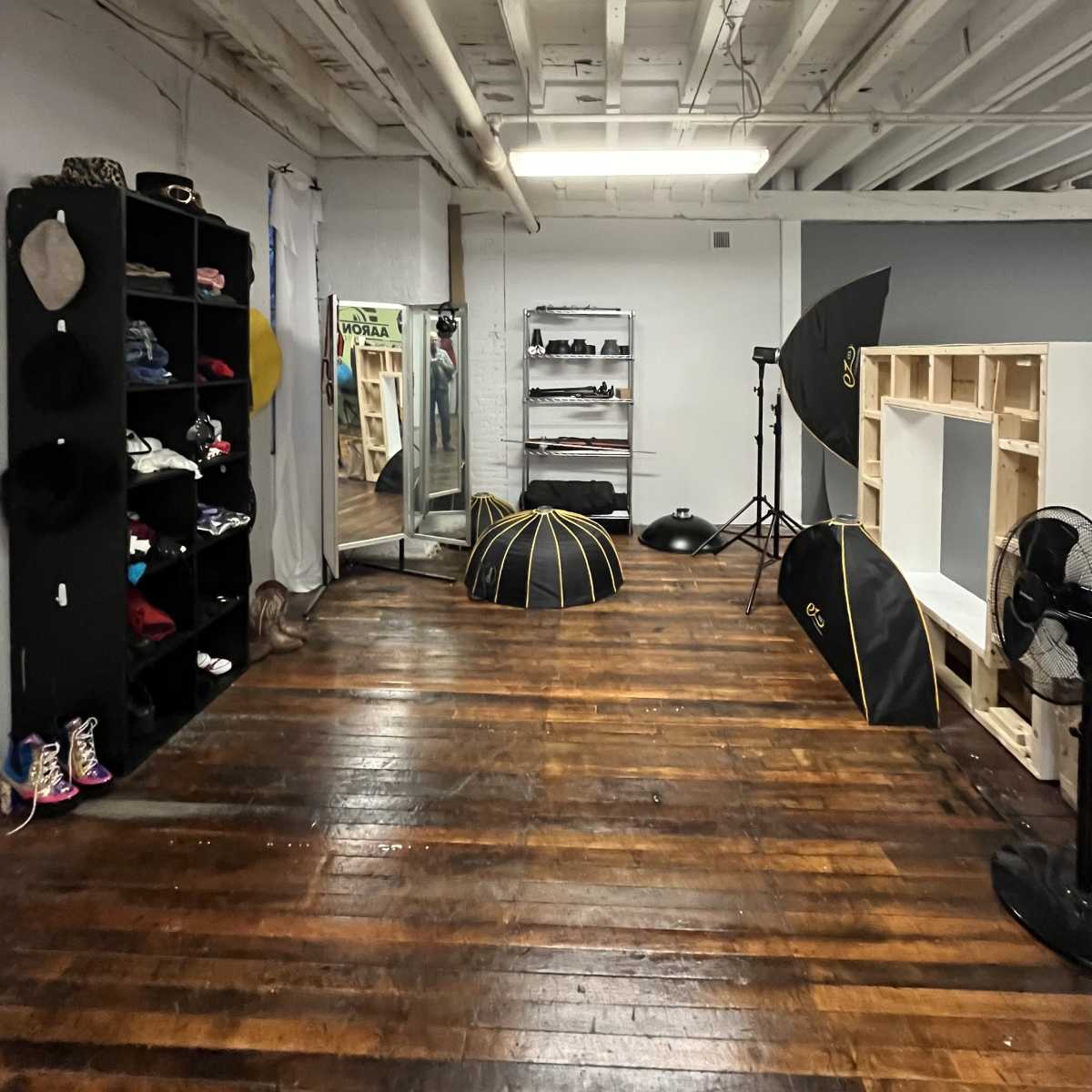 A spacious room with wooden floors, featuring a clothing rack, shelves filled with clothes, a full-length mirror, and photography equipment including light umbrellas.