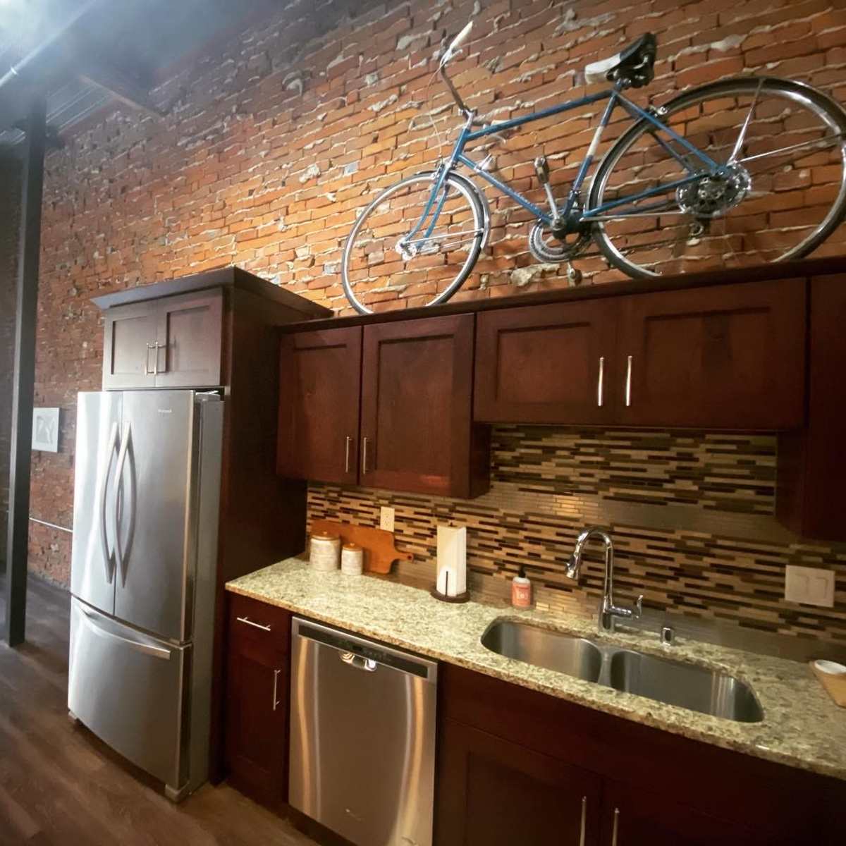 A modern kitchen with wood cabinets, a granite countertop, and a bicycle mounted on a brick wall.