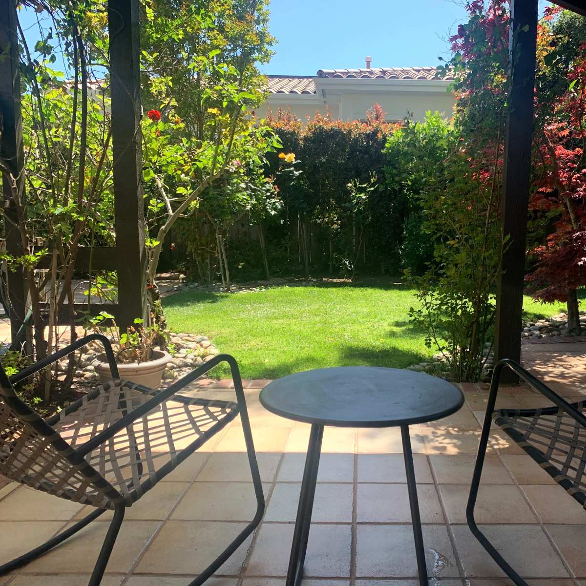 A shaded patio area features two chairs and a small table overlooking a green lawn surrounded by various plants and trees.