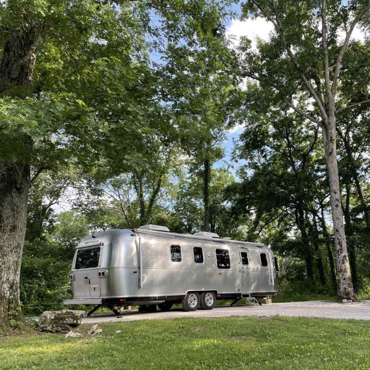 A silver Airstream trailer is parked on a gravel site surrounded by tall green trees.