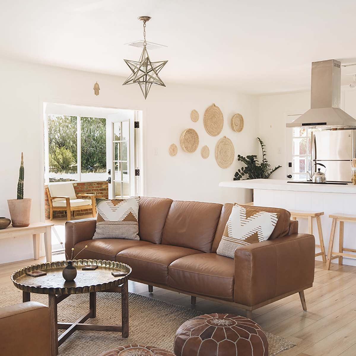 A modern living room with a brown leather sofa, a round coffee table, and a dining area visible through an open door, accented by plants and decorative wall hangings.