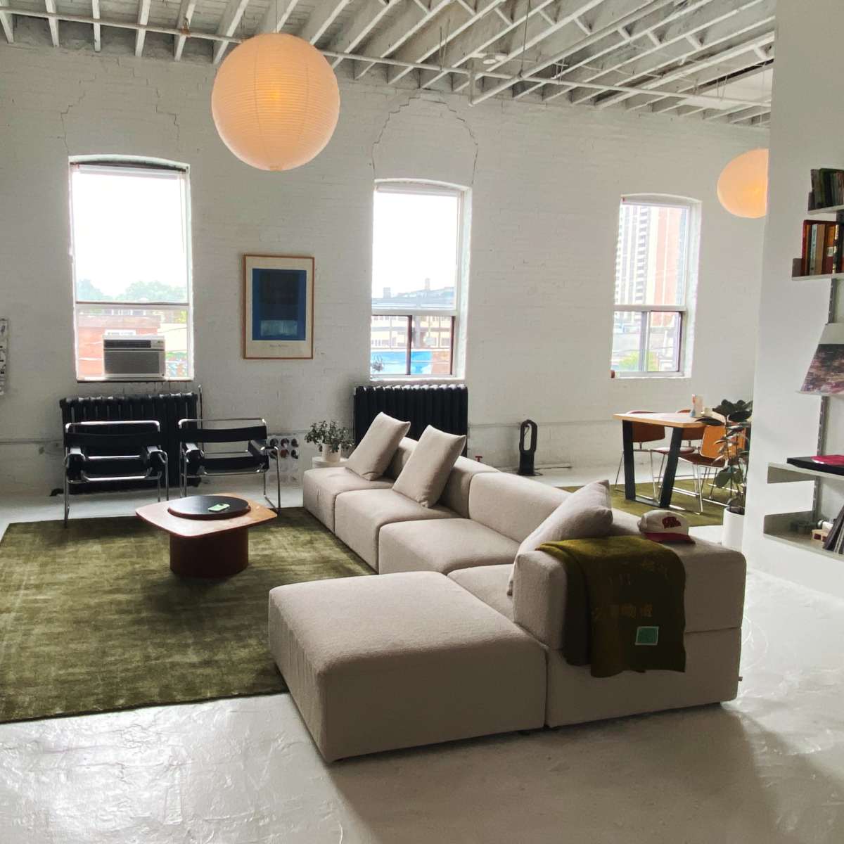 An open-plan living space features a sectional sofa, a round coffee table, and large windows allowing natural light, with a dining area visible in the background.