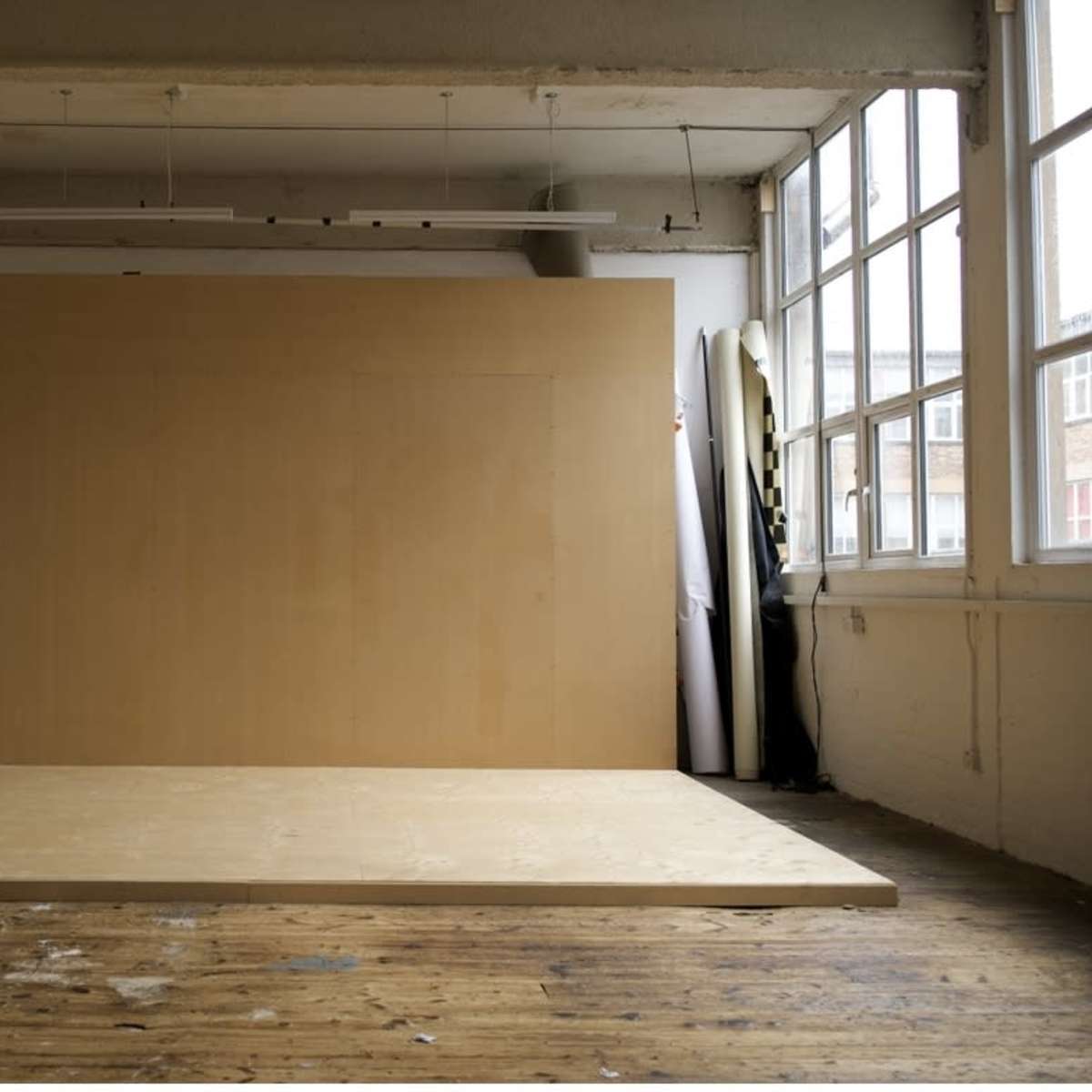 A spacious photo studio with a wooden platform and a large blank wall, illuminated by natural light from the windows.