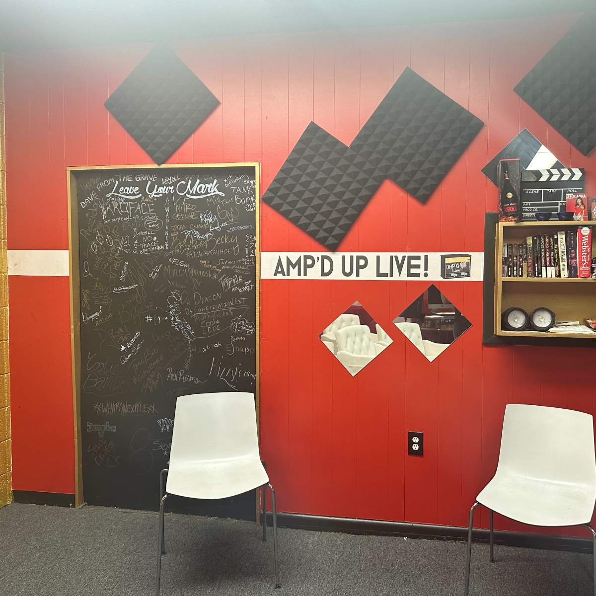 A room with red walls, two white chairs, a black chalkboard covered in writing, acoustic foam panels, and a shelf displaying various items.