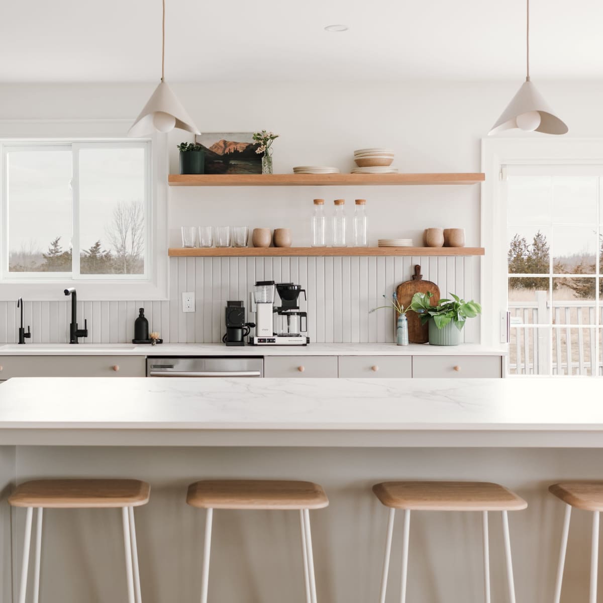 A modern kitchen features a central island with three wooden stools, open shelving with dishware, and a black faucet above a white countertop.
