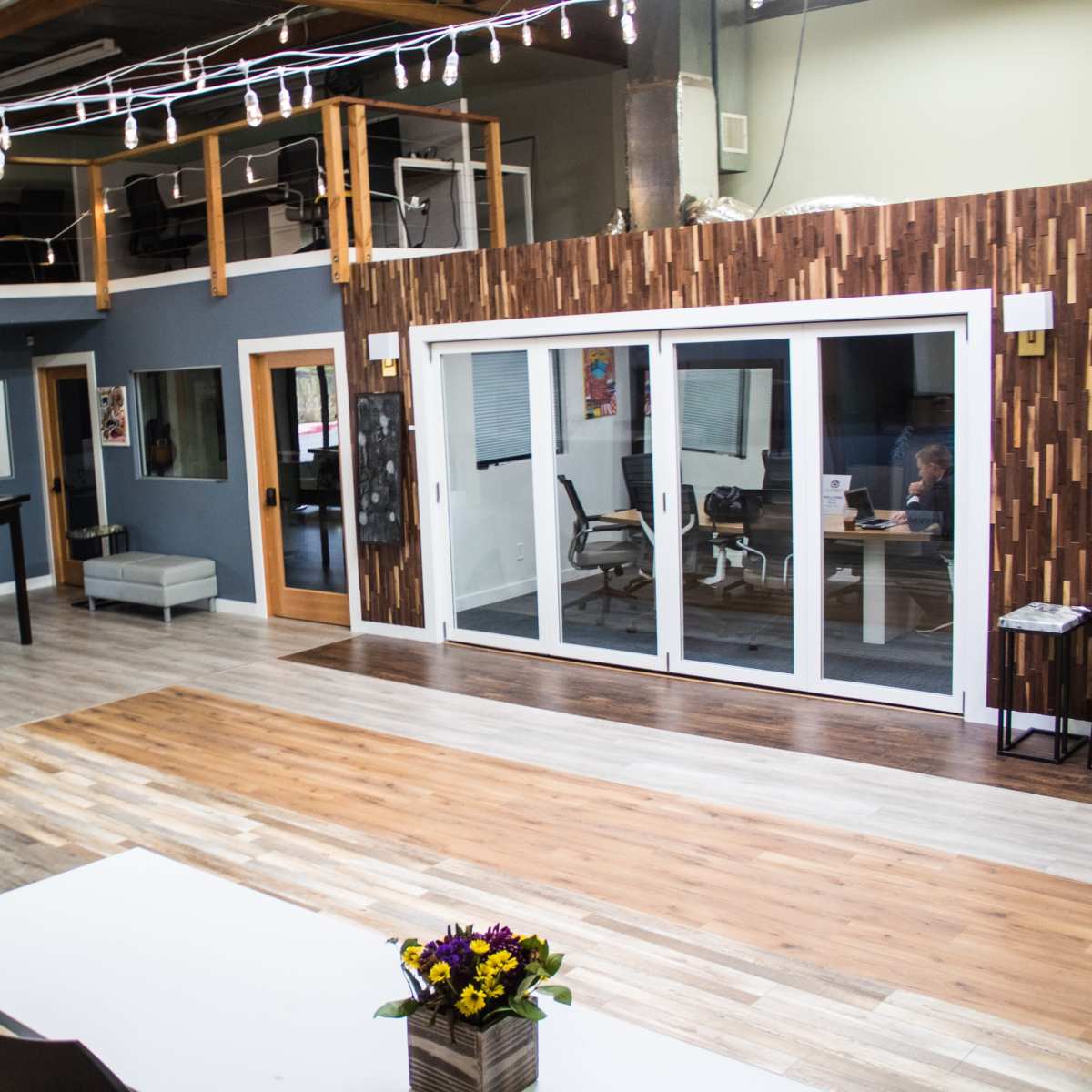 A spacious, modern interior with wooden flooring, multiple doors leading to separate rooms, and a large glass door area that opens to a brightly lit workspace.