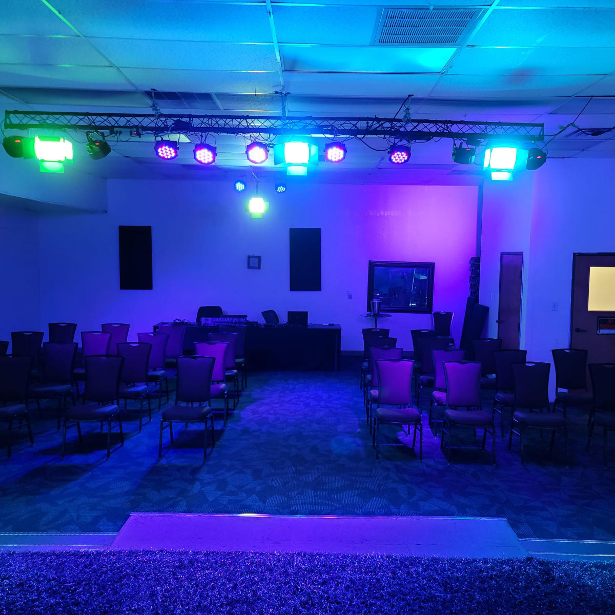 A dimly lit event space with rows of chairs facing a stage, equipped with colorful lights and sound equipment.
