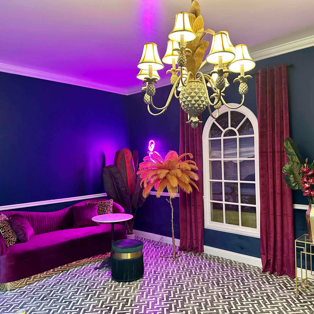 A stylish lounge features a purple velvet sofa, a round table, and an ornate chandelier against dark blue walls, complemented by a decorative window and potted plants.
