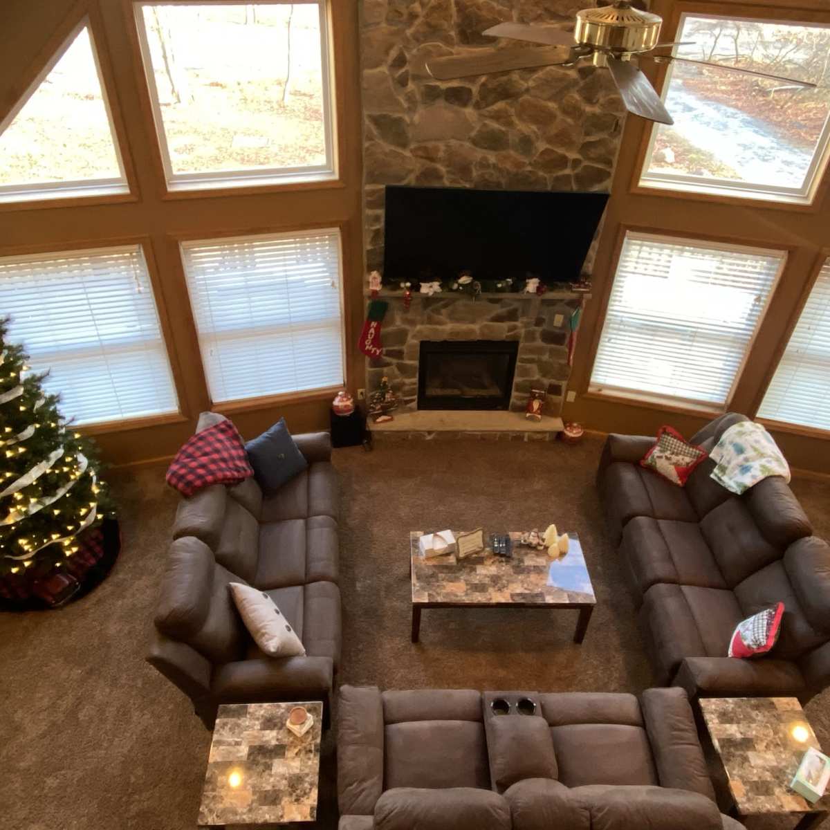 A spacious living room with a stone fireplace, a large Christmas tree decorated with lights, and two couches arranged around a coffee table.