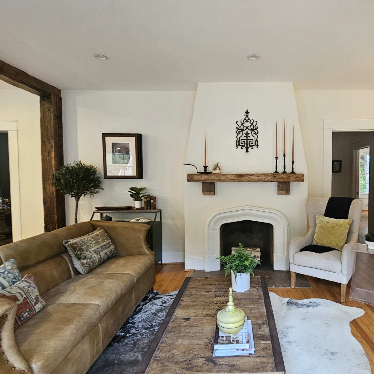 A cozy living room features a brown sofa, a wooden coffee table, and a white fireplace flanked by candles and plants.