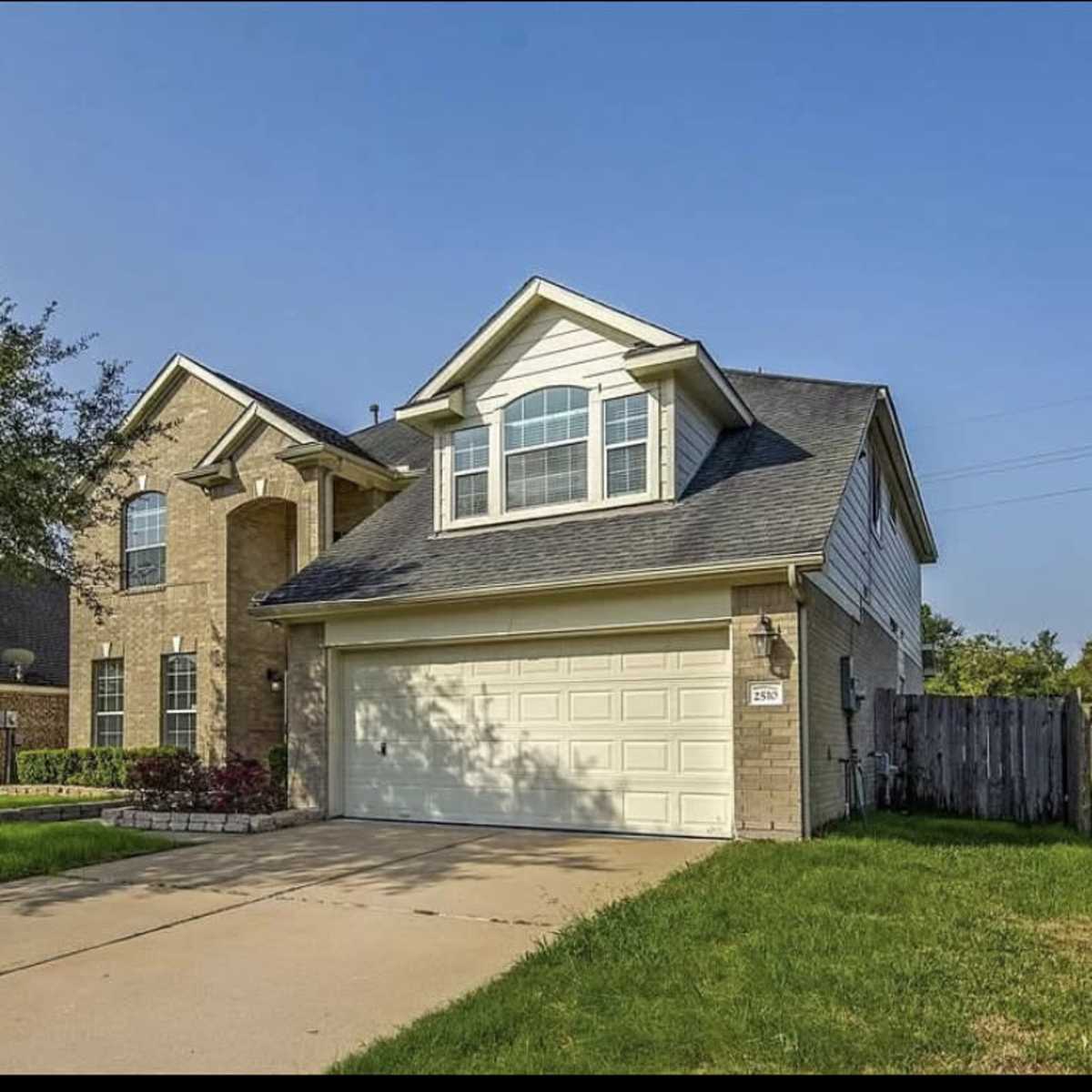 A two-story brick house with a gable roof and a two-car garage is situated on a grassy lot.